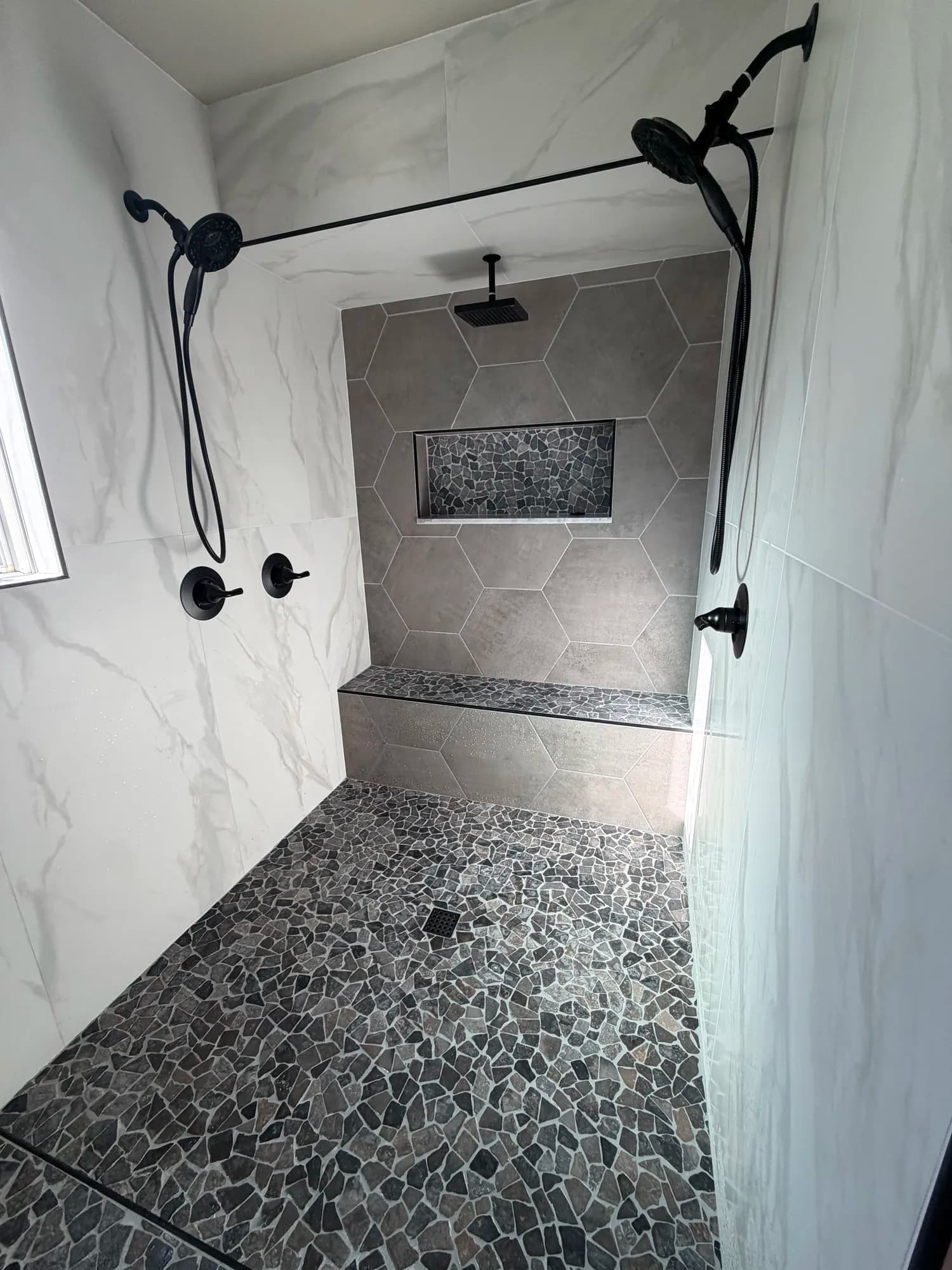 Curbless walk-in shower with gray hexagon tile accent wall, pebble mosaic floor and bench, and dual matte black shower heads in Menifee home
